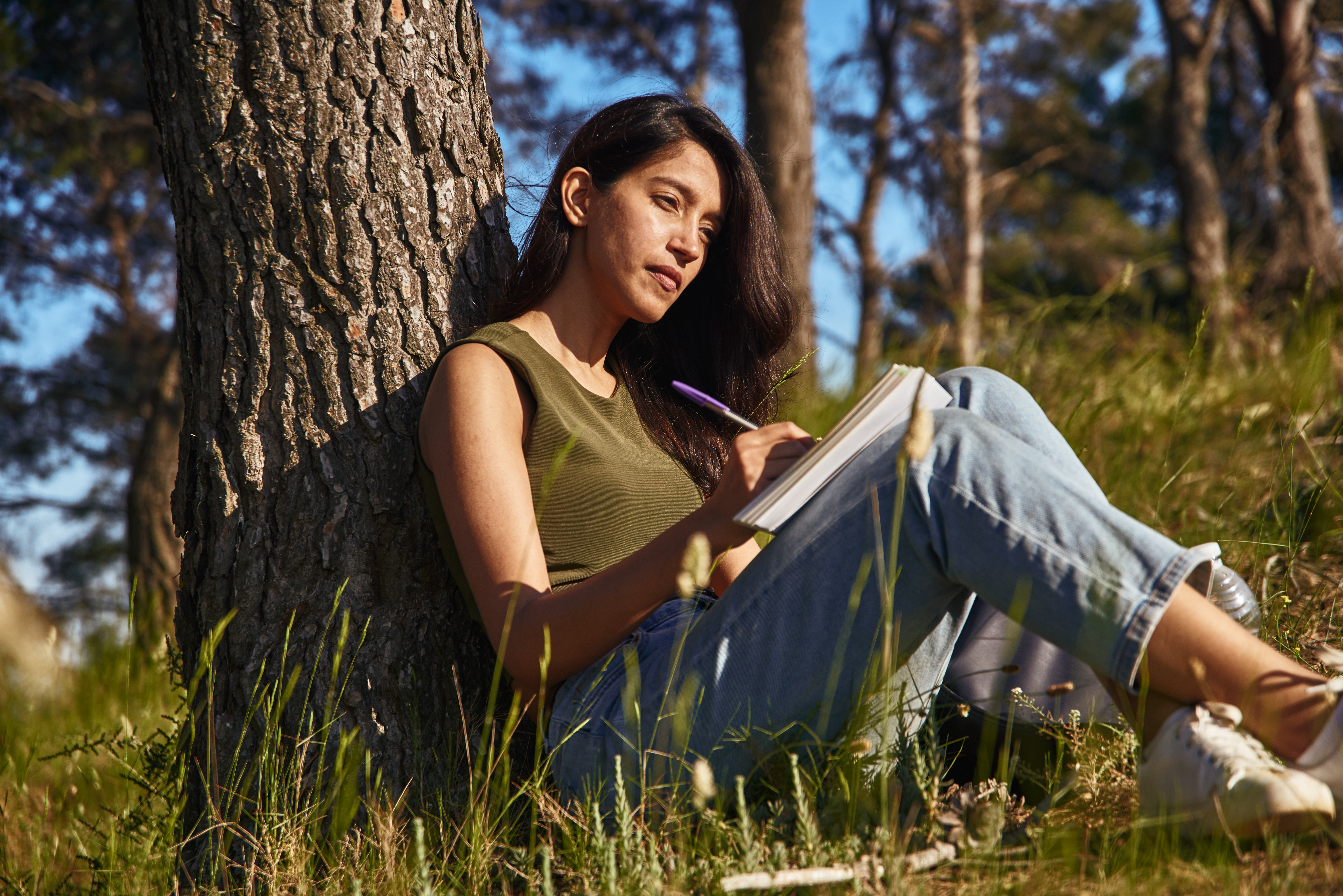 A young woman with dark hair sits at the base of a tree in a forested area. She is holding in her lap a journal and pen. 
