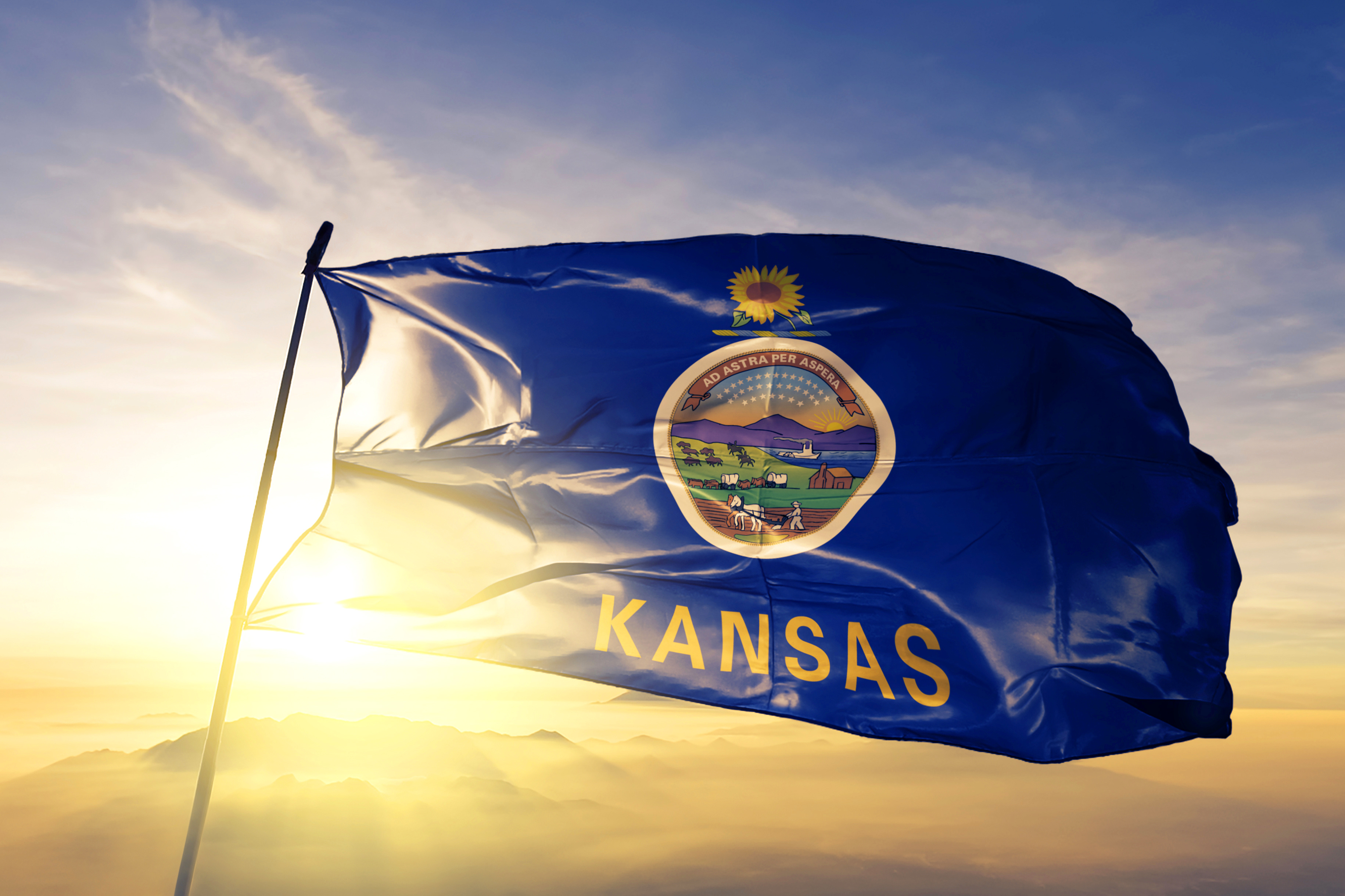 The Kansas state flag blows in the wind while the sun sets behind it across a cloud-streaked sky. 
