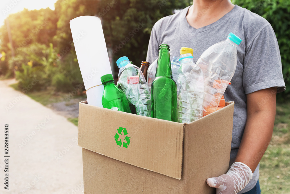 A person wearing gloves is holding a cardboard box with lots of plastic items in it. The symbol for "reduce, reuse, recycle" is on the front of the box. 