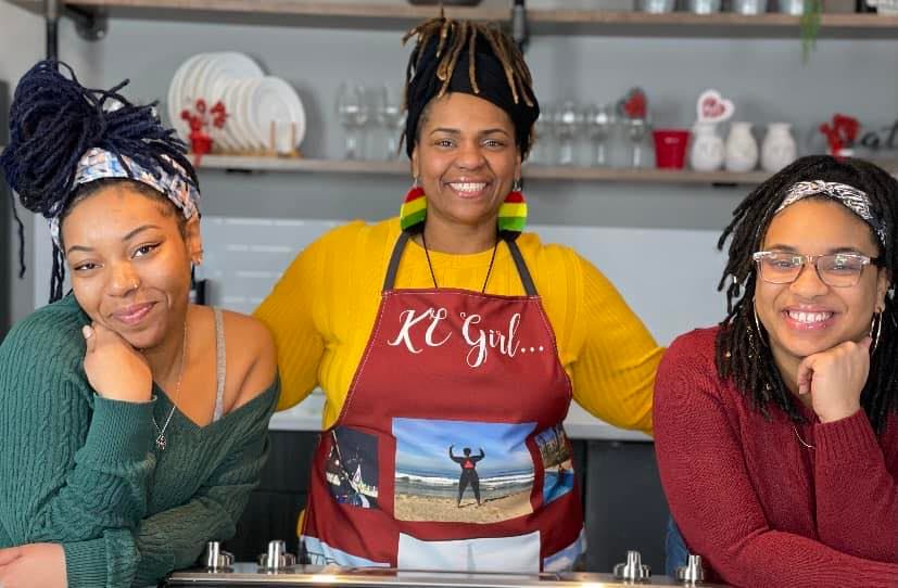 Three women with dark-skin and culturally Black hairstyles stand in a kitchen. The middle woman, Stacey Hardman, wears a red apron that says "KC Girl" on it. 