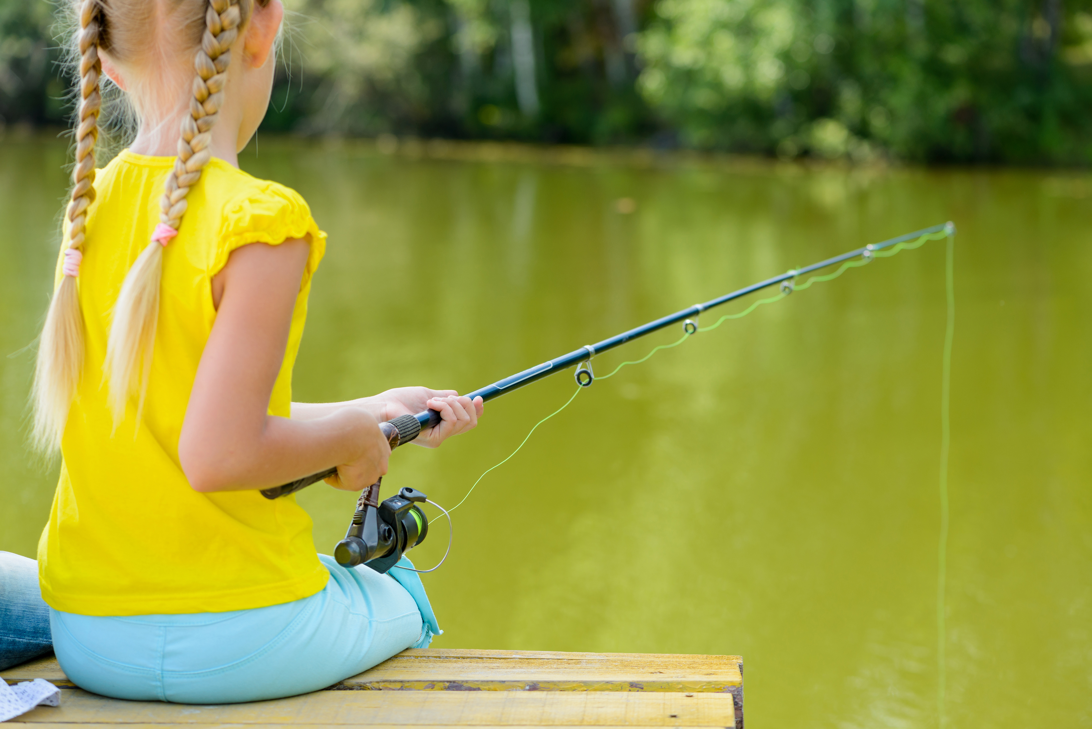 A young, blonde child with two pigtails sits on a dock made of wood while holding a fishing pole. 