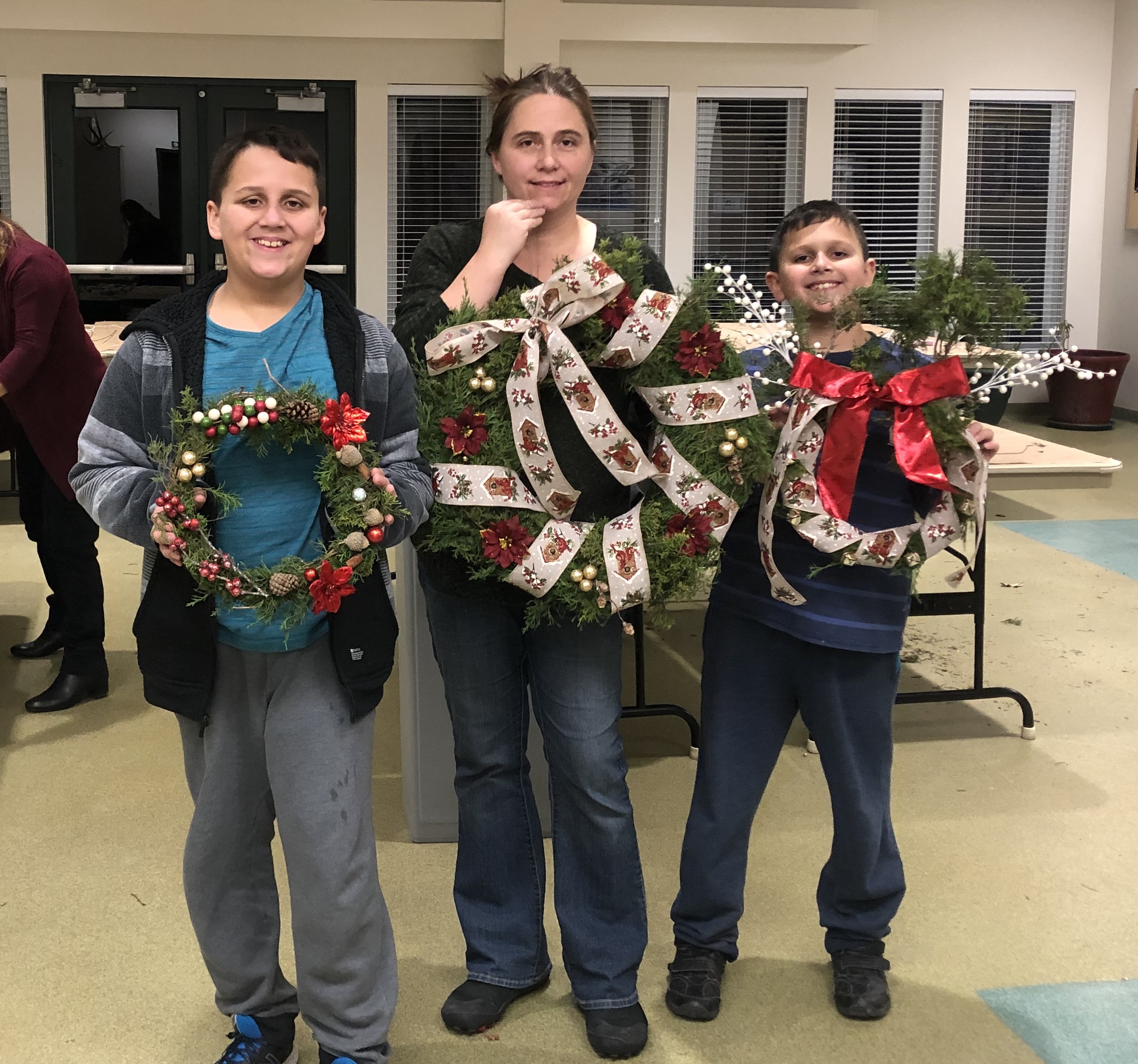 Two children stand next to their parent holding holiday wreaths. 