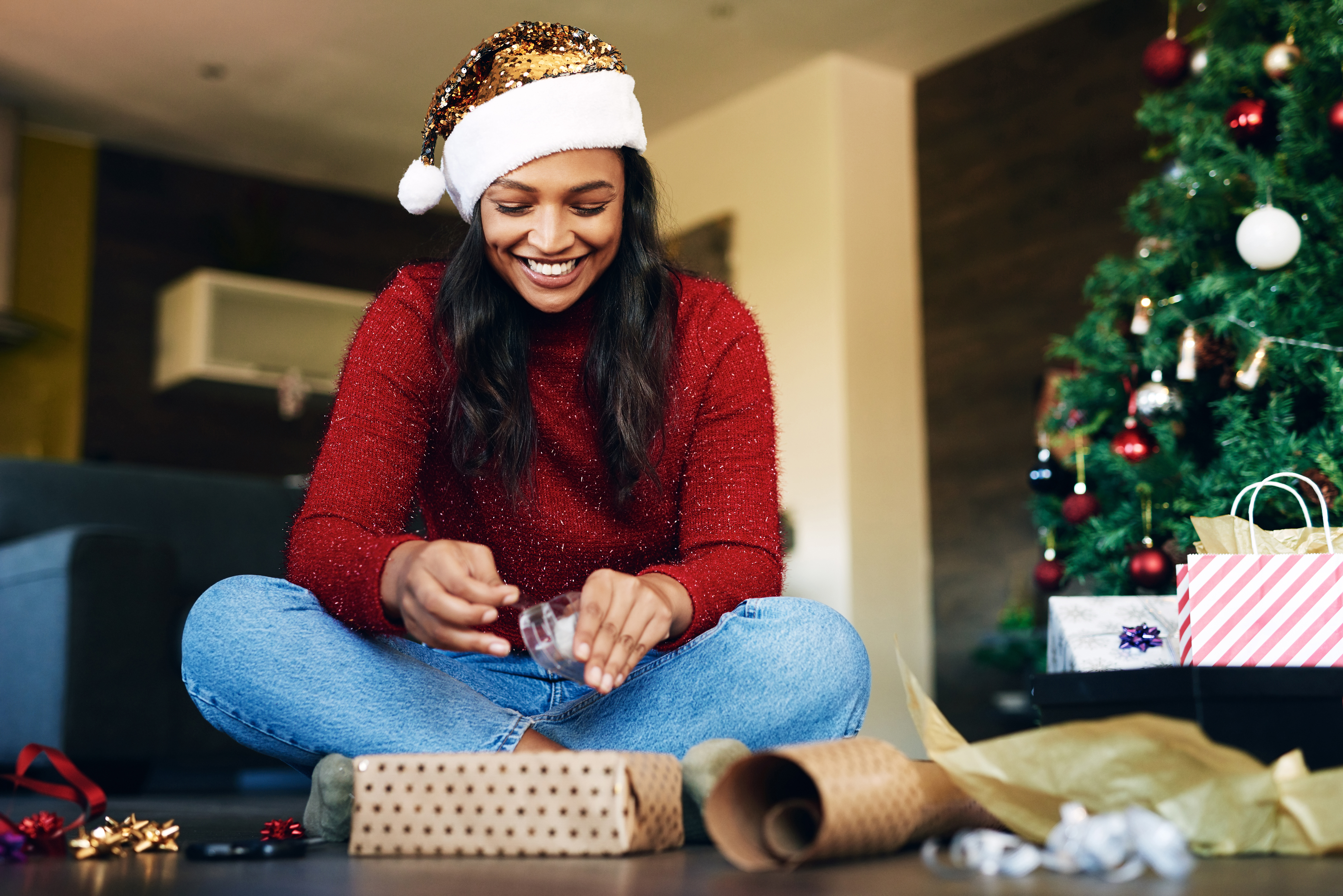 Black woman in a sparkly gold santa claus hat sitting on the floor wrapping presents with a smile on her face.