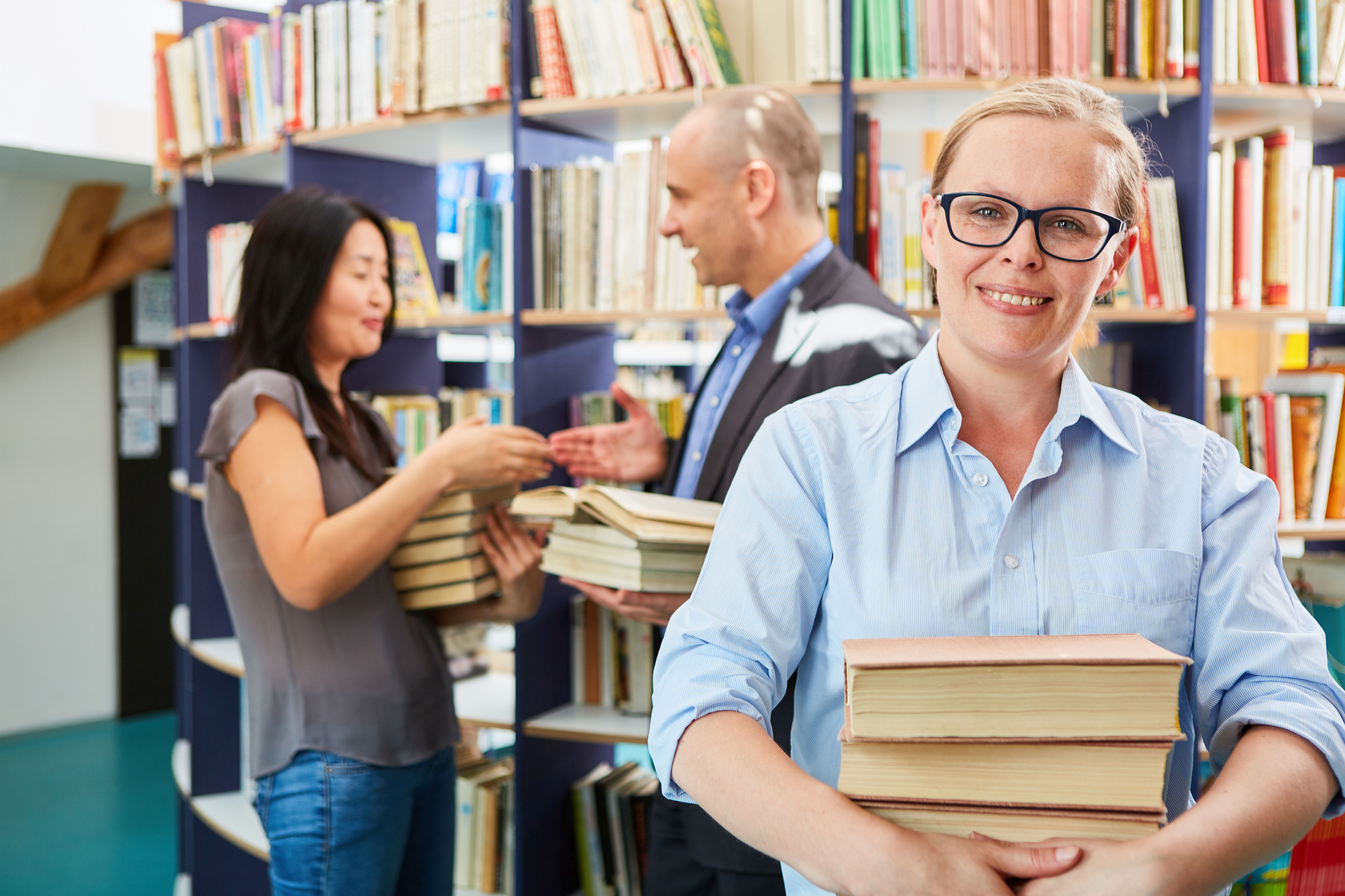 Person standing in foreground smiling with a stack of books in hand. Two people in background also holding books and shaking hands.