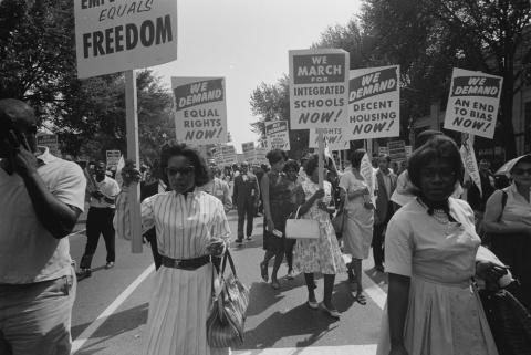 Photograph shows a procession of African Americans carrying signs for equal rights, integrated schools, decent housing, and an end to bias.