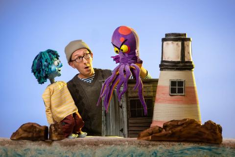 Puppeteer holding two puppets on a set with a lighthouse. 