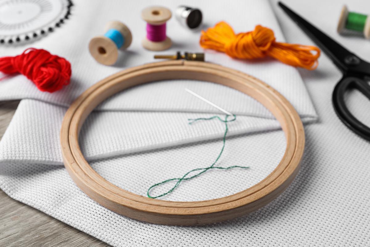 Supplies for needlepoint including wooden embroidery hoop, thread in various colors, black scissors, white fabric, and an embroidery needle with green thread.